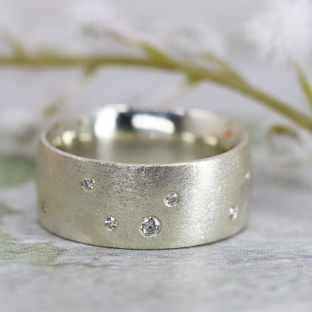 A wide silver ring with small diamonds set into the surface. There is a green twig above the ring