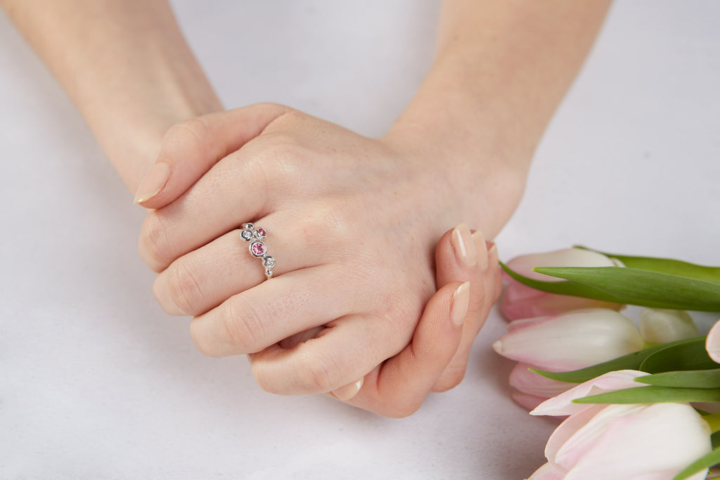 A pair of hands clasped next to a bunch of pink tulips. On one finger is a silver ring with pink tourmaline gemstones