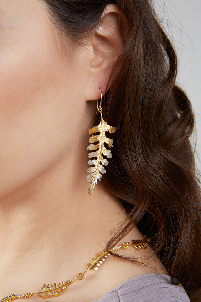 The side of a woman's face with dark brown long hair. Her ear is exposed. A large silver and gold fern shaped earring hangs from the ear with a silver wire going through the ear