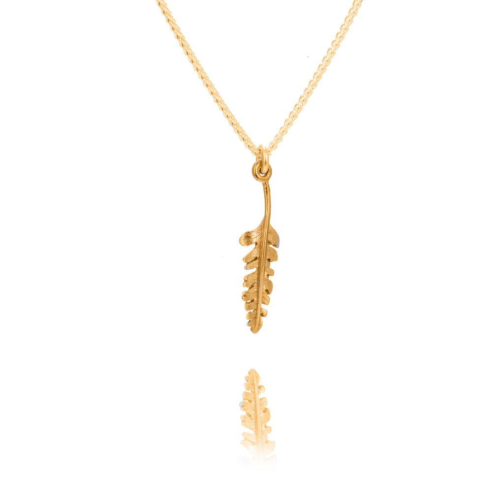 A tiny fern shaped gold pendant on a gold chain. There is a reflection of the fern tip undernaeth