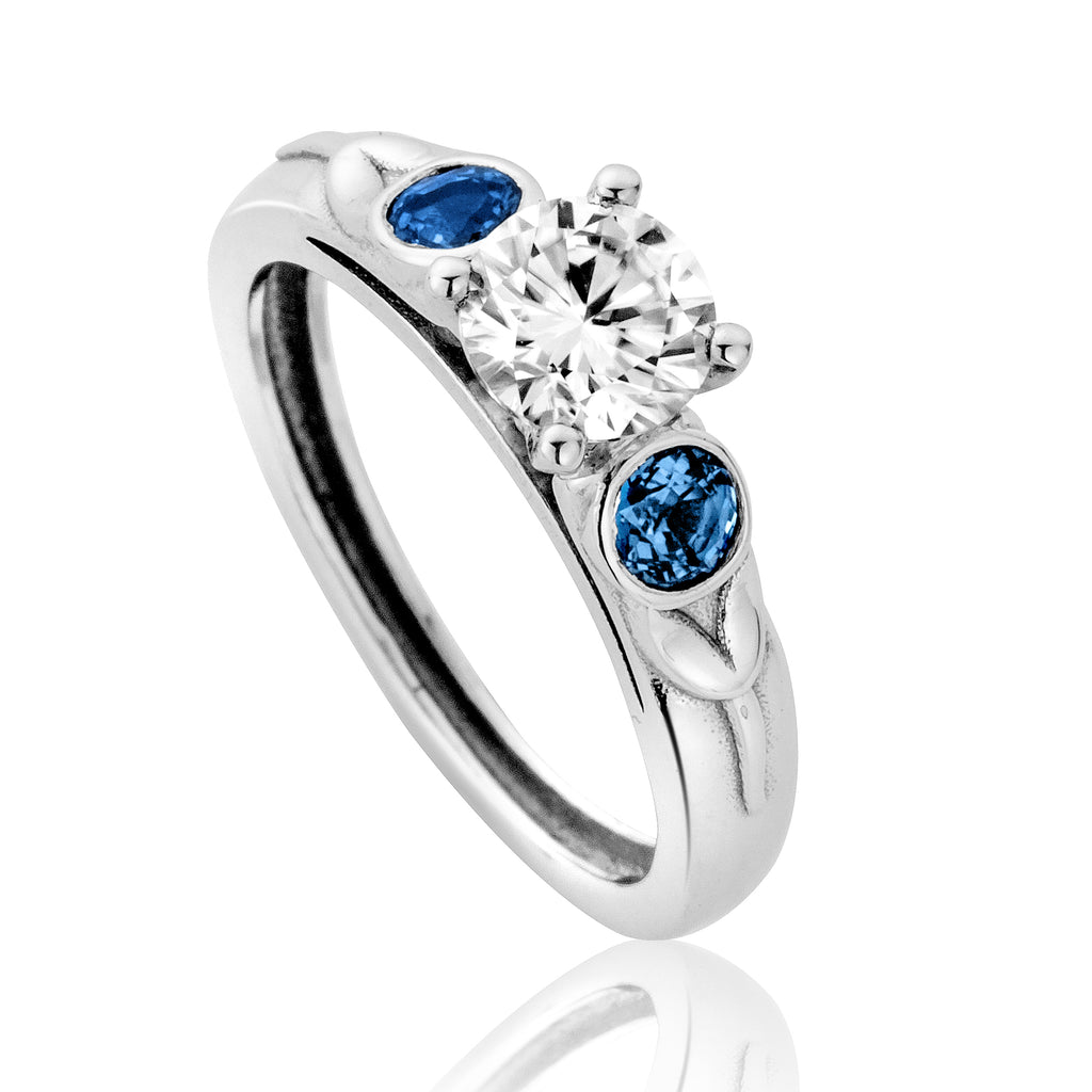White gold engagement ring with leaf pattern on shoulders, a white central stone and 2 blue stones