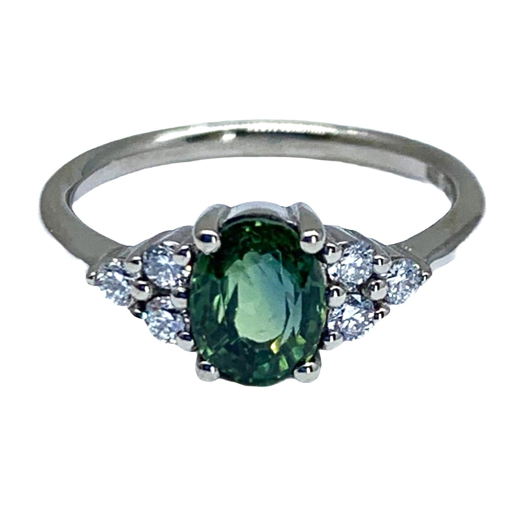 A white gold ring lying on its side with an oval green stone at the front. At each side of the green stone are 3 clear stones in a triangular fomation