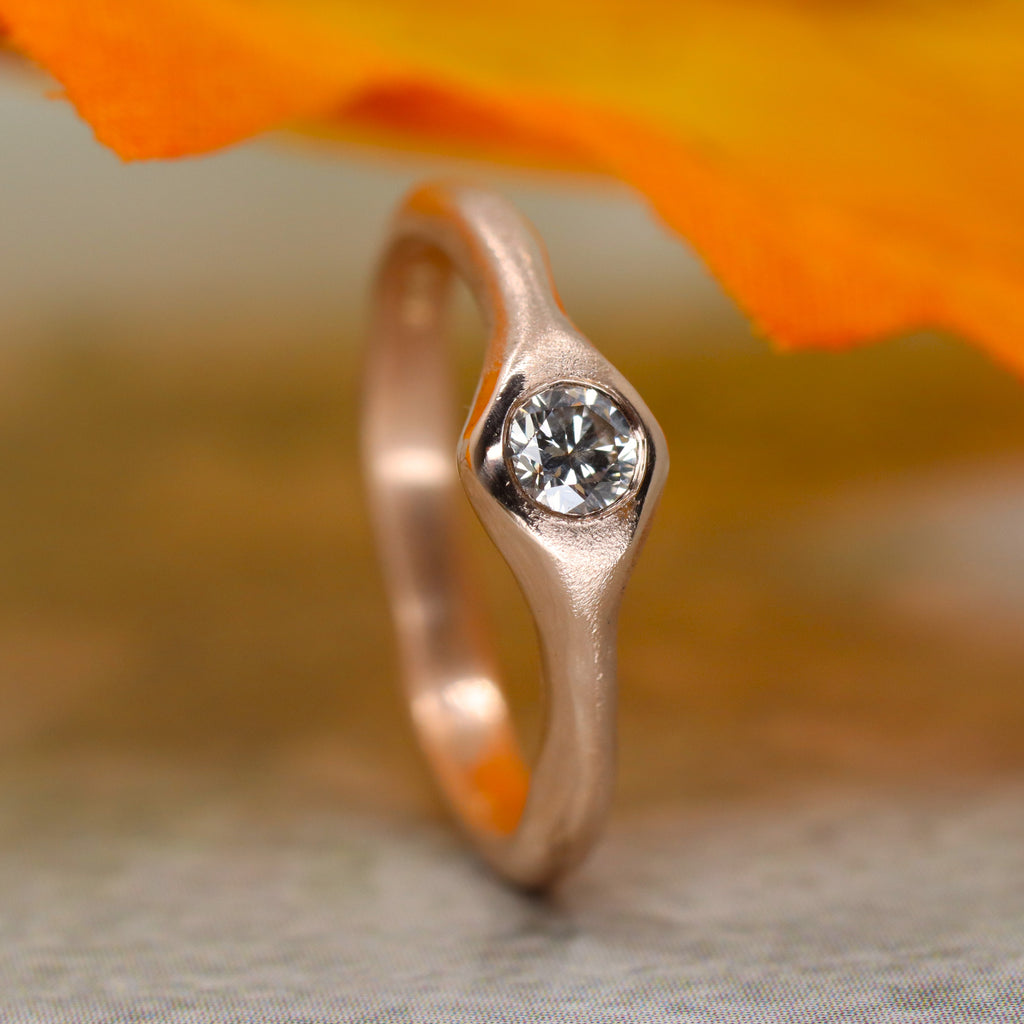 A wavey rose gold ring standing on end. The ring widens out at the front and a round diamond is set flush with the surface. Above the ring is an orange leaf