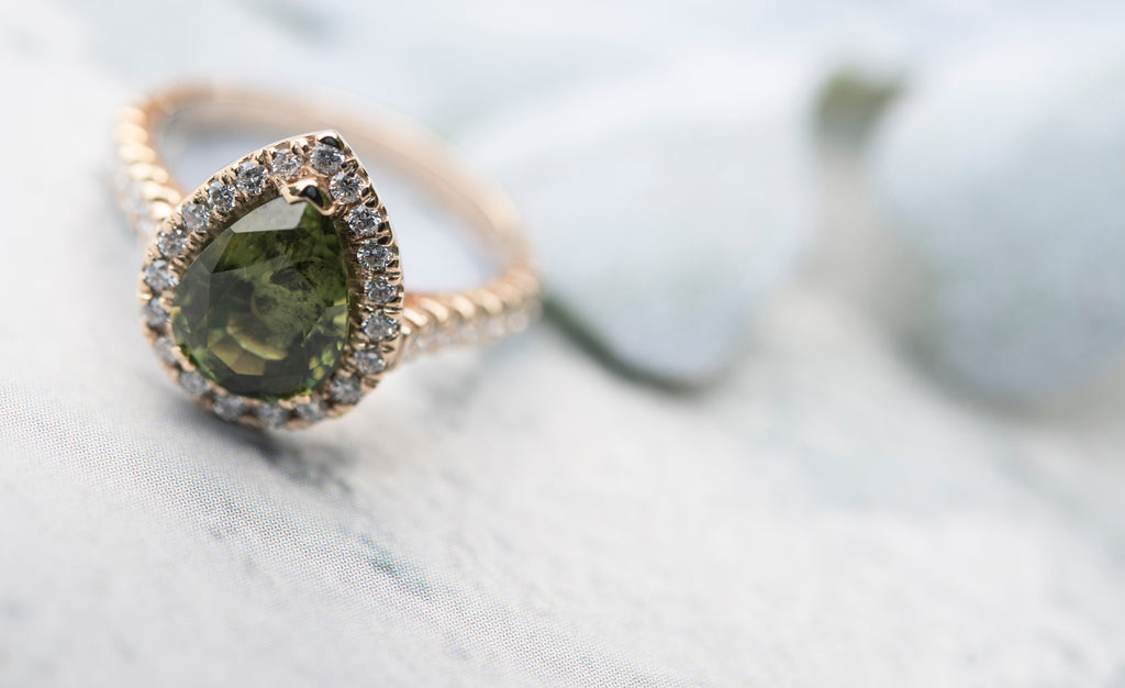 Yellow gold pear shaped halo engagement ring with a one of a kind pear shaped green sapphire surrounded by diamonds and with diamond set shoulders
