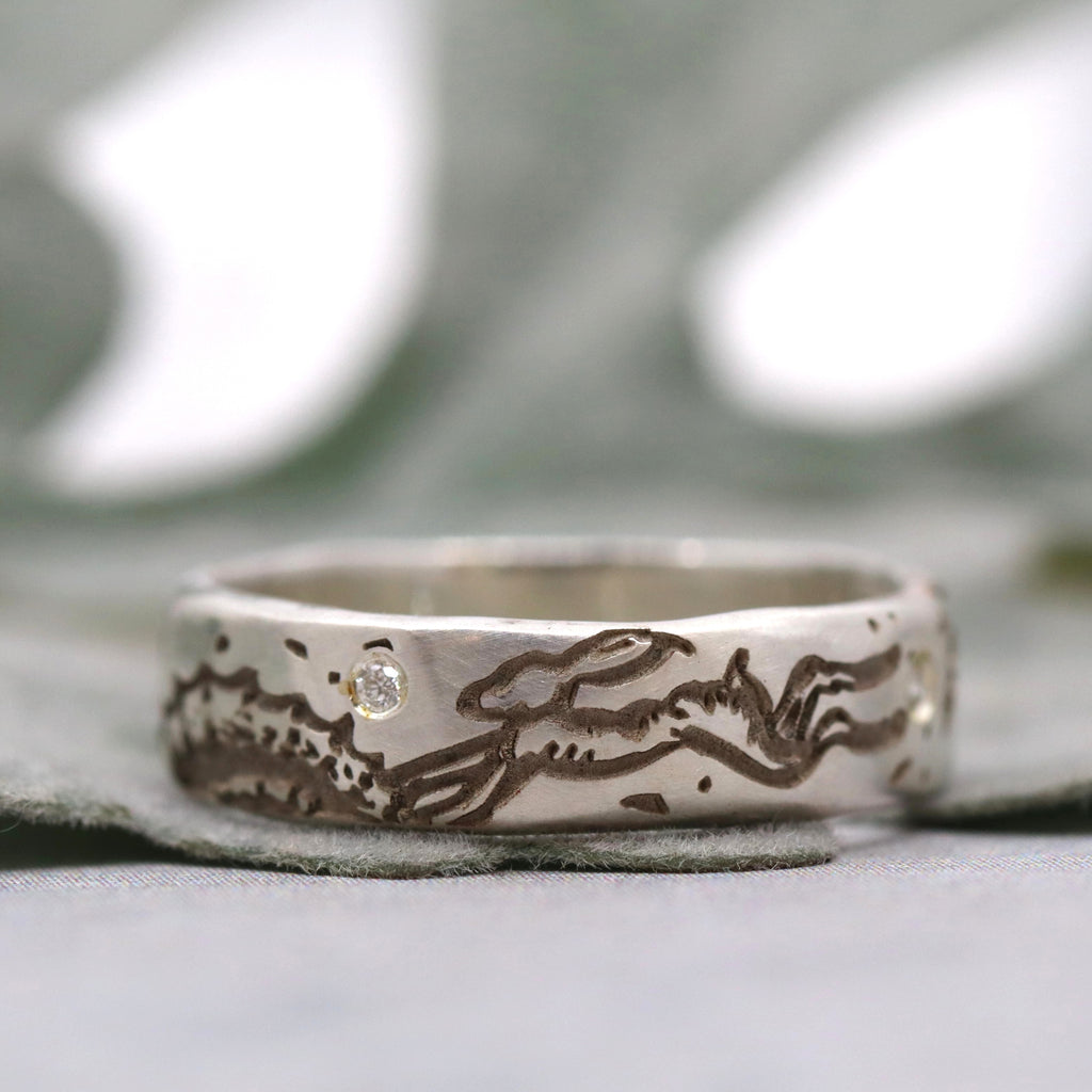 Silver ring with engraved hare design and small diamond on a blurred background