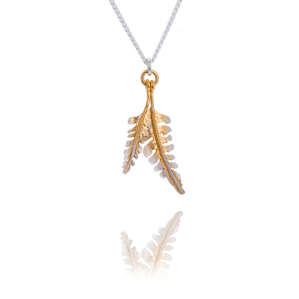 A tiny and larger fern shaped pendant hanging from a gold circle. The gold circle is attached to a silver chain. The ferns are gold down the centre with silver edges. There is a reflection of the tips of the pendant underneath