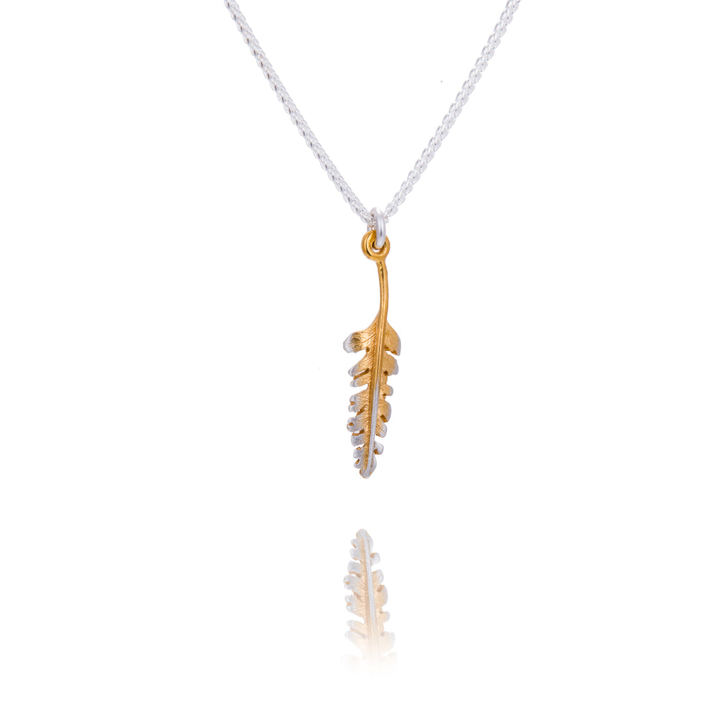 A tiny fern shaped pendant with gold down the centre and silver edges. The pendant is hanging from a silver chain. There is a reflection of the pendant tip underneath