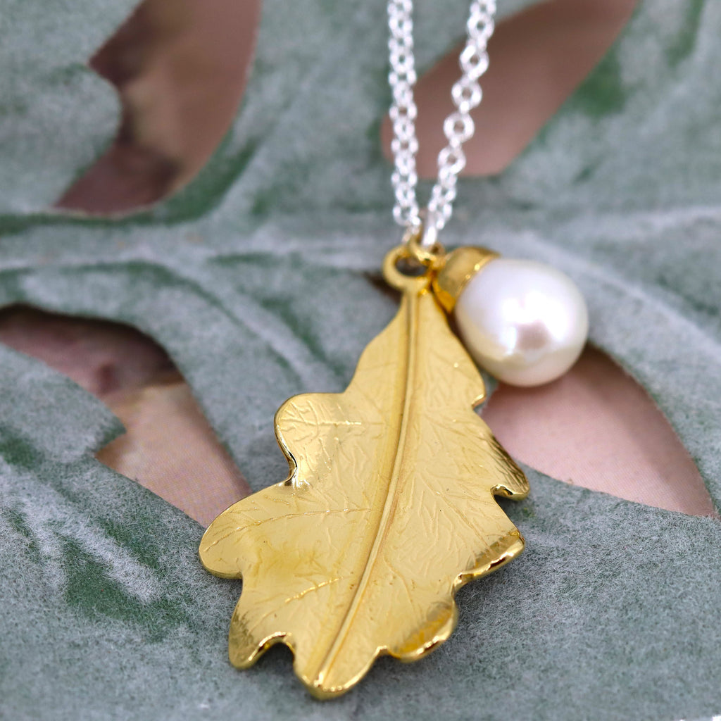A gold oak leaf pendant with white pearl on a silver chain. The necklace is lying on some green leaves.