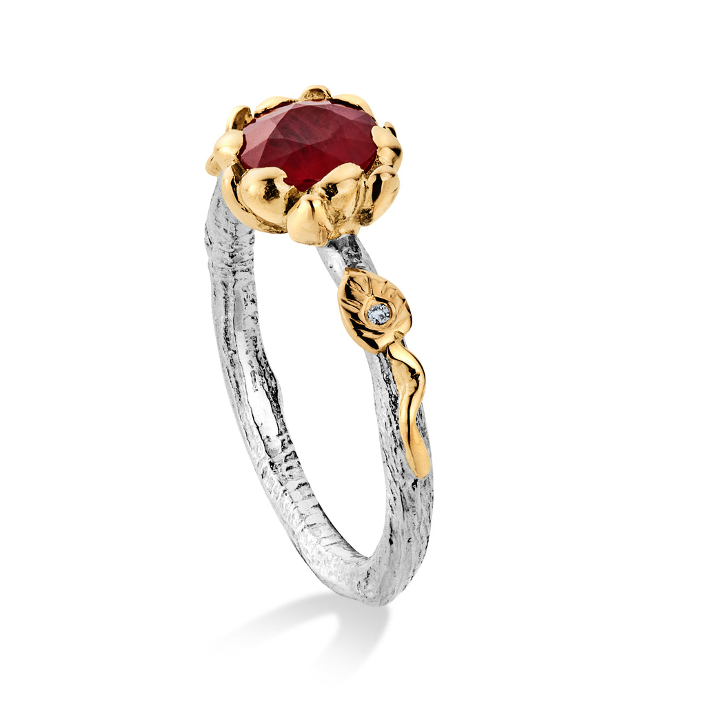 White gold engagement ring standing on end with a large ruby at the top in a yellow gold flower setting. There is a gold leaf and tendril on the shoulder set with a diamond.