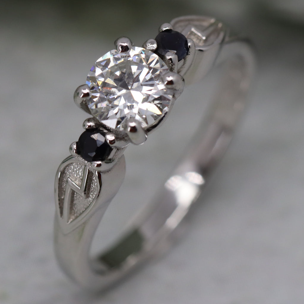Platinum engagement ring standing on end. There is a shield shape on each shoulder containing a Harry Potter style lightning bolt. Between the shields is a large round white diamond and two smaller black sapphires