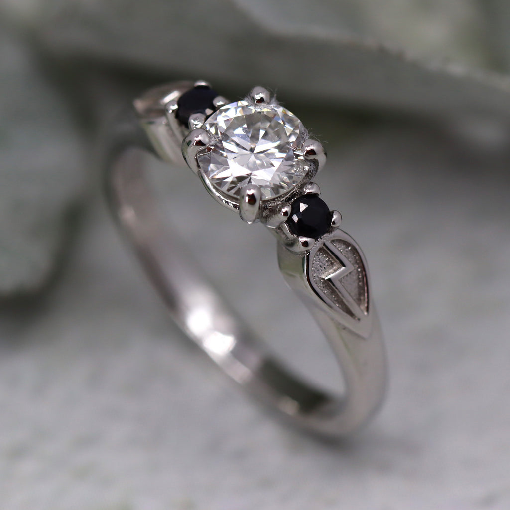 White gold engagement ring standing on end with leaves in the background. At the top of the ring a round diamond is set in claws with a black stone at either side of the diamond. Below the black stone on the right side is a sheild shape containing a lightning bolt