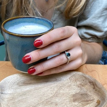 A hand with painted red nails holding a blue cup. There is a green gemstone ring on the second finger of the hand.