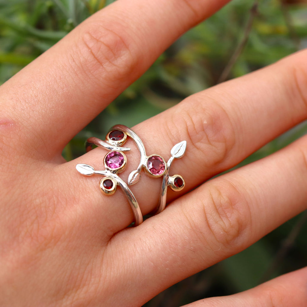 Two delicate silver vine shaped rings with pink and red stones being worn on the middle finger of a hand