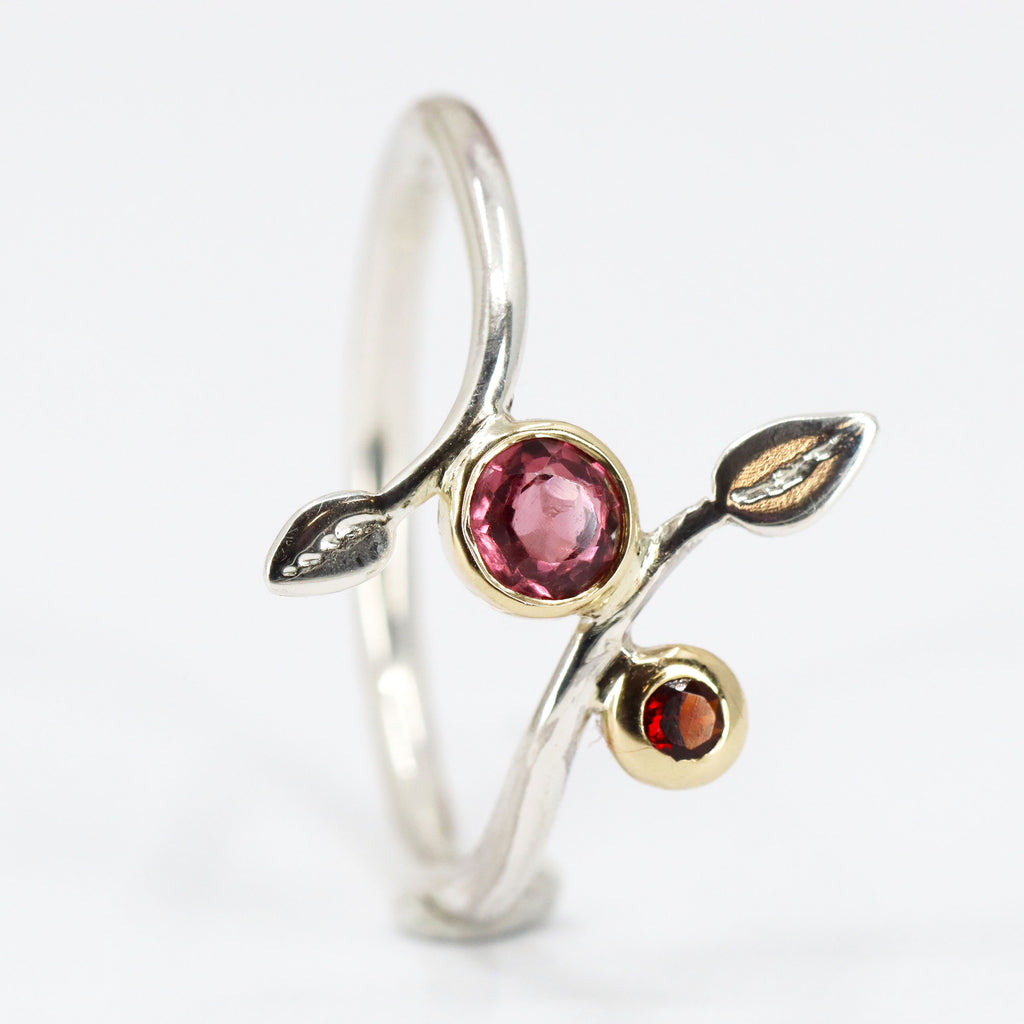 A silver wire ring standing on end. On each end of the silver wire that makes the ring is a silver leaf. A pink stone is set in between the leaves and a red stone is set off to the side