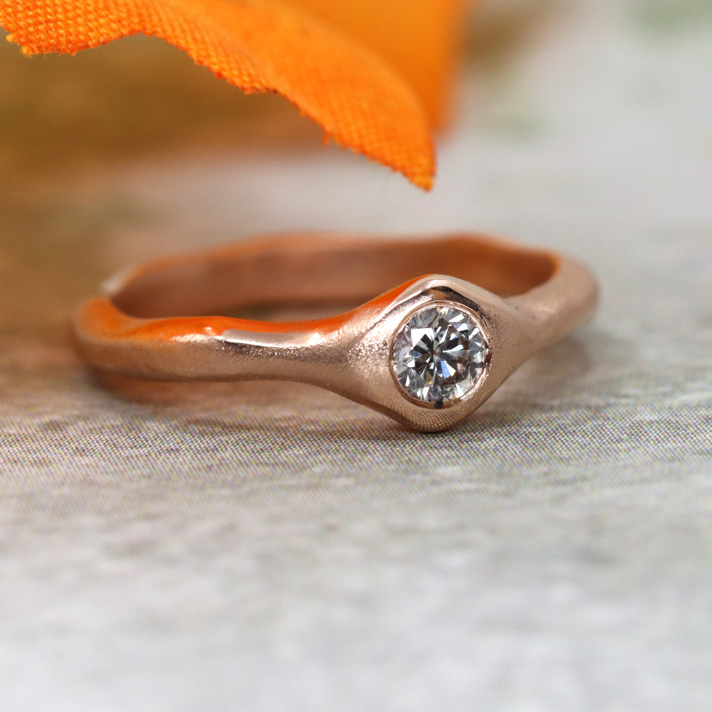 A rose gold ring with wavey sides on a grey cloth. The ring widens out to a peak on each side. A diamond is set in the widest part. There is an orange leaf above the ring
