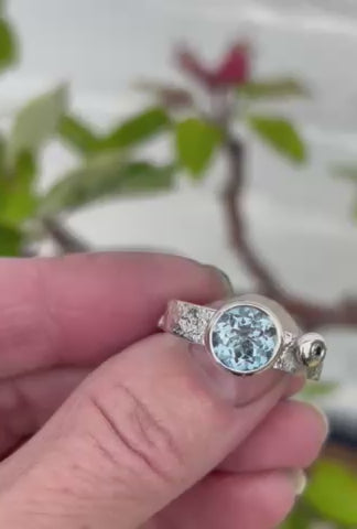 A silver ring with light blue stone being moved around in a hand, with a tree in the background