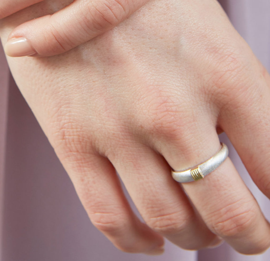 A hand wearing a matt silver ring with 5 shiny gold wires on the top