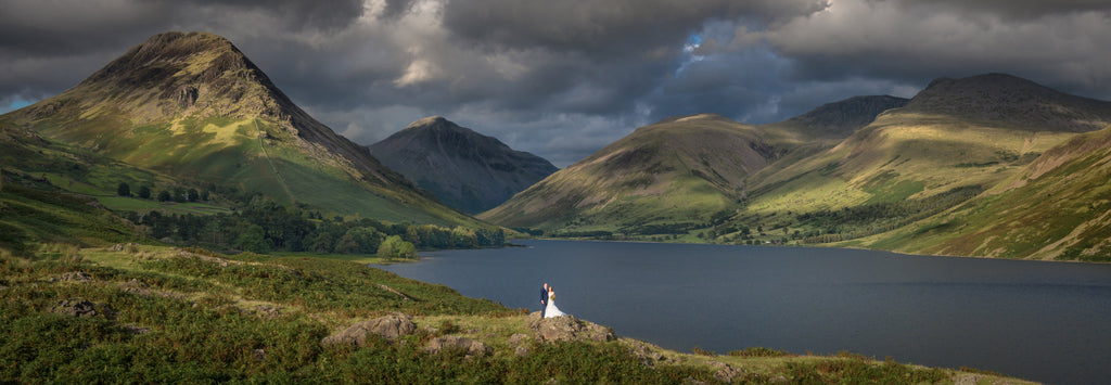 A dramatic landscape with a bridal couple standing next to a lake, surrounded by hills in the Lake District