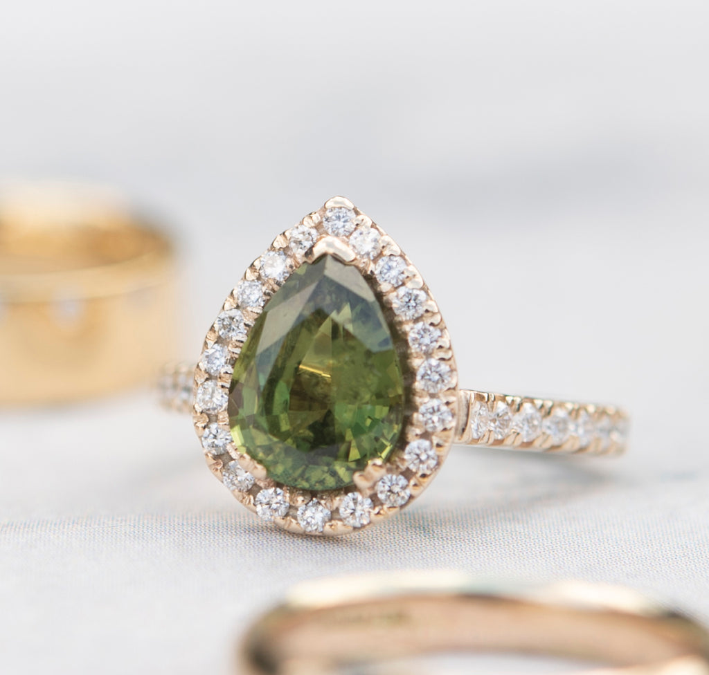 Pear shaped green sapphire diamond halo ring sitting amongst other gold rings