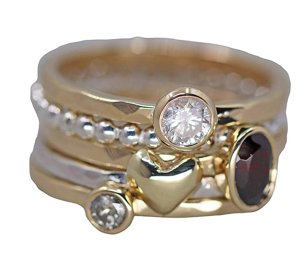 A stack of 3 gold and 2 silver rings, one with an oval garnet at the front, 2 with round diamonds at the front and 1 with a gold heart at the front.