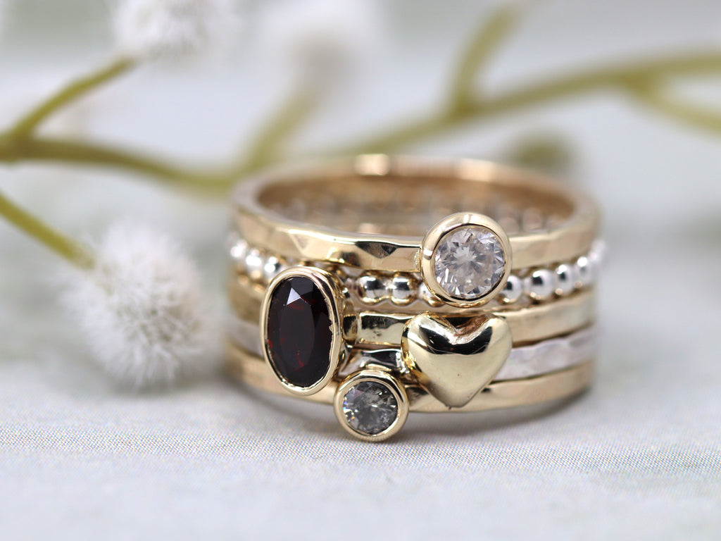 A stack of 5 rings. Top to bottom - hammered gold ring with round diamond, beaded silver ring, hammered gold ring with oval garnet, hammered silver ring with gold heart, hammered gold ring with round diamond