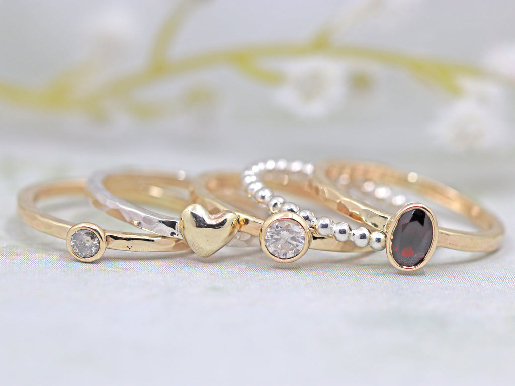 A set of rings made from recycled gold and gemstones. Left to right - hammered gold ring with small round diamond, hammered silver ring with gold heart, hammered gold ring with large round diamond, beaded silver ring, hammered gold ring with oval garnet