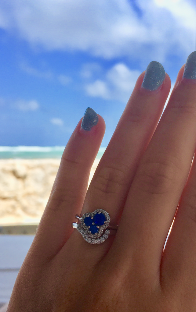 A hand with blue fingernails wearing a 3 blue sapphire engagement ring with diamond halo in front of a bright blue sky